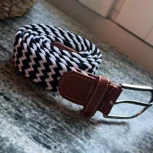 Svartvitt flätat bälte med skinnspänne - Snyggt flätat bälte i svart och vitt med bruna skinn-detaljer vid spännet. Bältet har ett randigt mönster och ett klassiskt metallspänne. Perfekt accessoar för att lyfta din outfit med en cool kontrast.