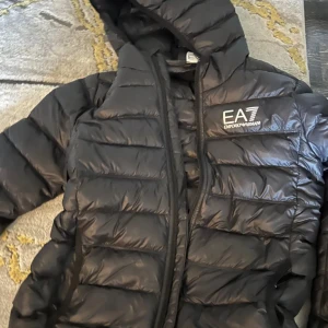 Svart pufferjacka från Emporio Armani - Säljer en svart pufferjacka från Emporio Armani EA7 med huva och dragkedja. Jackan har ett stilrent, quiltat mönster och logga på bröstet. Perfekt för kyliga dagar och ger en sportig vibe. Passar dig som gillar snygga och funktionella ytterplagg.