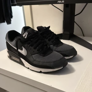 Nike Air Max 90 svart och grå - Snygga Nike Air Max 90 sneakers i svart och grå med vita detaljer. Skorna har klassisk siluett, synlig Air Max-dämpning i sulan och mesh- samt skinnpaneler för en sportig look. Perfekta för dig som gillar streetstyle och vill ha bekväma sneakers med ikonisk design.