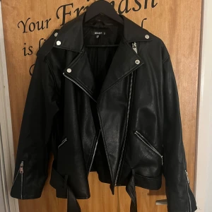 Svart skinnjacka från Missguided - Snygg svart skinnjacka i bikerstil från Missguided. Jackan har silvriga dragkedjor, breda slag, bälte i midjan och detaljerade knappar på axlarna. 