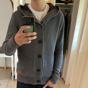 Stickad kofta från Rocklin - Mysig grå kofta med knappar från Rocklin med stora knappar, huva och två fickor framtill. Insidan är fodrad med svart fleece för extra värme. Perfekt för kyliga dagar och har en snygg, enkel design med ribbade muddar och nederkant.