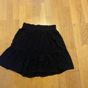 Svart volangkjol med resårmidja - En svart kortkjol med volang och bred resår i midjan. Kjolen har en luftig och lätt känsla, perfekt för dig som gillar en enkel men trendig stil. Passar till många olika toppar och sneakers.