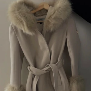 Hollies kappa - Snygg beige kappa från Hollies med stora fuskpälsdetaljer på huva och ärmslut. Kappan har omlottknäppning och bälte i midjan för en clean och elegant look. Perfekt för kalla dagar när du vill vara både varm och stilren.