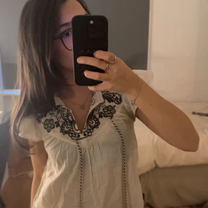 Vit blus med broderi och knytband - Supersöt vit blus med svarta broderade blommor runt v-ringningen och knytband med tofsar framtill. Blusen har korta ärmar och en luftig, lite lös passform. Perfekt för dig som gillar boho-vibbar och detaljer.