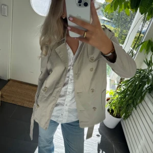 Beige trenchcoat med bälte - Snygg beige trenchcoat med dubbelknäppning och bälte i midjan. Jackan har klassisk krage, långa ärmar och två framfickor. Perfekt för dig som gillar stilrena och tidlösa plagg. Passar till jeans och sneakers för en chill look.