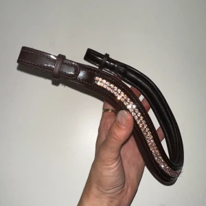 Brunt pannband med glittriga stenar. Full - Säljer ett mörkbrunt, glossy pannband i skinn med glittriga stenar i rosé. Går att knäppa på snabbt med kardborrefästen i sidorna. Helt oanvänt! Storlek Full. 