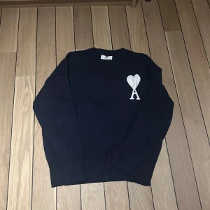 Ami Paris stickad tröja - Säljer en mörkblå stickad tröja från Ami Paris med ikoniskt vitt hjärta och A på bröstet. Tröjan har rund hals, ribbade muddar och är långärmad. Perfekt för dig som gillar stilrena och trendiga plagg. Hör av er vid eventuella funderingar! 😁🤝🏼