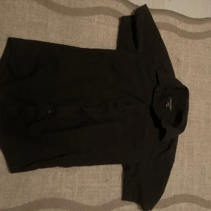 Svart kortärmad skjorta från George -                                Snabb leverans.                                Svart skjorta från George med klassisk krage och korta ärmar. Skjortan har knappar framtill och är tillverkad i en mix av bomull och polyester, vilket gör den både mjuk och lätt. Perfekt för dig som gillar en enkel och clean stil.