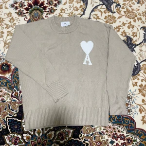 Beige stickad tröja från Ami Paris - Säljer en beige stickad tröja från Ami Paris med deras ikoniska vita hjärta och A-logga på bröstet. Tröjan har rund hals, ribbade muddar och är tillverkad i mjuk bomull. Perfekt för dig som gillar stilrena och trendiga plagg.