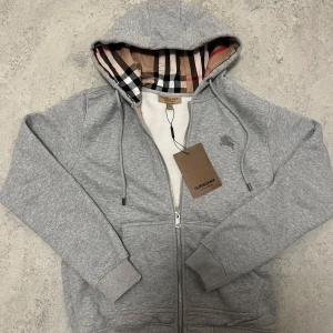 Grå hoodie från Burberry med dragkedja - Snygg grå hoodie från Burberry med klassiskt rutigt foder i huvan och broderad logga på bröstet. Tröjan har dragkedja framtill, snörning i huvan och ribbade muddar. Perfekt för dig som gillar stilrena och exklusiva plagg.