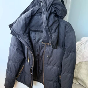 Svart dunjacka från InWear med huva - Svart dunjacka från InWear med avtagbar huva och snygga guldiga dragkedjor. Jackan är quiltad och har flera fickor med dragkedja. Perfekt för kalla dagar och har en klassisk, lite puffig look.