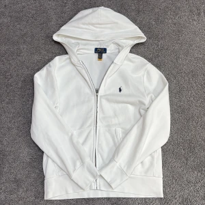 Vit hoodie från Polo Ralph Lauren - Vit hoodie från Polo Ralph Lauren med dragkedja och klassisk blå logga broderad på bröstet. Tröjan har huva, fickor framtill och är tillverkad i mjuk bomull. Perfekt för en clean och stilren look.