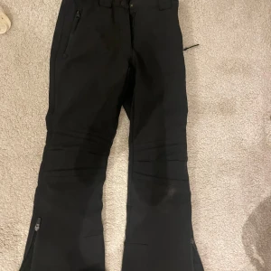 Svarta täckbyxor från McKinley stl 146 - Svarta täckbyxor från McKinley i storlek 146. Byxorna har dragkedjor vid bensluten och praktiska fickor med dragkedja. Perfekta för vinterns alla äventyr och håller dig varm och torr i snön.