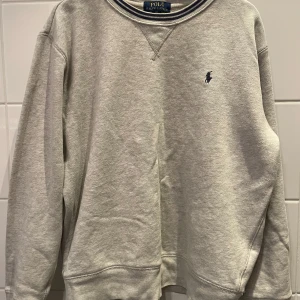 Grå sweatshirt från Polo Ralph Lauren - Klassisk grå sweatshirt från Polo Ralph Lauren med rund halsringning och broderad marinblå logga på bröstet. Tröjan har långa ärmar och ribbade muddar vid ärmslut och nederkant. Perfekt för en chill och stilren look.