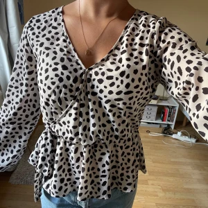 Prickig omlottblus med knyt i midjan - Snygg vit blus med svarta prickar och omlottmodell. Blusen har v-ringning, långa ärmar med smock vid ärmslut och ett knytband i midjan som ger en fin siluett. Tillverkad i ett lätt och luftigt material, perfekt för en trendig look.