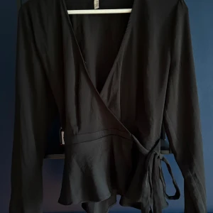 Svart omlottblus från NLY Trend - Säljer en svart omlottblus från NLY Trend i storlek 34. Blusen har långa ärmar, knytband i midjan och en snygg v-ringning. Perfekt för dig som gillar stilrena och trendiga plagg.