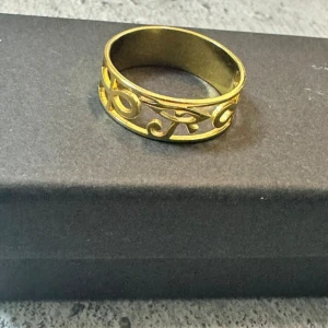 Guldfärgad ring med ögat av Horus och Ankh - Cool guldfärgad ring med ett öppet, utskuret mönster som visar det egyptiska ögat av Horus, Ankh och dekorativa snirkliga detaljer. Ringen har en bred design och glänsande finish som verkligen sticker ut på fingret. Steinless steel som tappar aldrig färg 