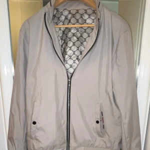 Beige vindjacka från Burberry - Snygg beige vindjacka från Burberry med klassiskt monogramfoder i svart och vitt. Jackan har dragkedja framtill, två fickor med knappdetaljer och diskreta ränder i rött och svart vid muddar och krage. Perfekt för dig som gillar stilrena och exklusiva plagg.