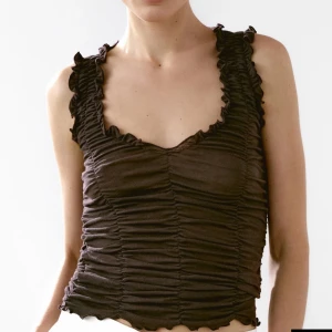 Brun rynkad linnetopp - Säljer en brun ärmlös linnetopp med rynkade detaljer och volangkant längs axelband och halsringning. Toppen har en croppad passform och är tillverkad i ett mjukt, stretchigt material som sitter snyggt på kroppen.