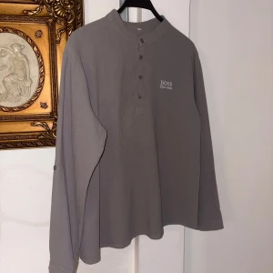 Grå långärmad pikétröja Hugo Boss - Snygg grå pikétröja från Hugo Boss med klassisk krage och knappslå framtill. Tröjan har lång ärm med knappdetalj och diskret broderad logga på bröstet. Perfekt för dig som gillar stilrena och enkla plagg med premiumkänsla. Använd 1 gång!