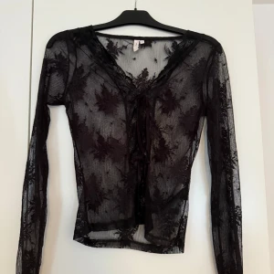 Svart meshblus med snörning från Nelly - Säljer en transparent svart meshblus från Nelly i storlek S. Blusen har långa ärmar, v-ringning och snygg snörning framtill. Blommiga spetsdetaljer ger en cool och trendig look. Perfekt att styla lager på lager eller med ett linne under.