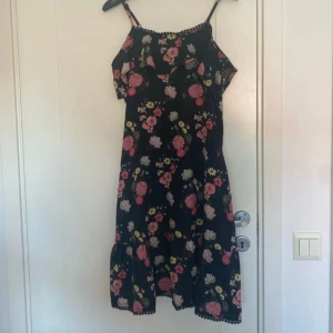 Svart blommig sommarklänning - Supersöt svart klänning med färgglada blommor i rosa, gult och vitt. Smala axelband (som är justerbara), volang upptill och dekorativ spetskant längs halsringningen. Lätt och flowy modell, perfekt för varma dagar. Klänningen är ärmlös och har en avslappnad passform. Lappen är avklippt, men uppskattad storlek är XS/S. 🤍