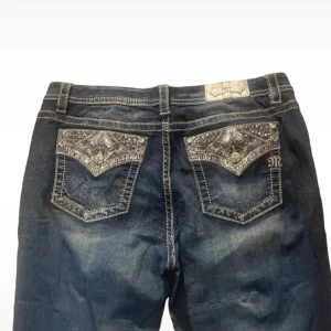 Mörkblå Miss Me jeans med blingfickor - Snygga mörkblå jeans från Miss Me med bootcut passform. Bakfickorna har blingade detaljer och broderier som verkligen sticker ut. Jeansen har kontrastsömmar och klassisk femficksdesign. Perfekta för dig som vill ha något extra. Storlek 34. Midja rakt över: 47cm Innerbenslängd: 77,5cm Midjehöjd: 25,5cm Benöppning: 24,5cm  Lite smutsiga där fram och en mindre lagning är gjord (syns på bild 2). Säljer då dom är för stora på mig.