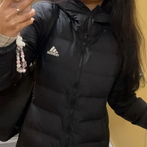 Svart pufferjacka från Adidas S - Säljer en svart pufferjacka från Adidas med huva och dragkedja framtill. Jackan har Adidas-loggan på bröstet. Perfekt för kyliga dagar och har en clean, sportig look. Jätte fin och varm!