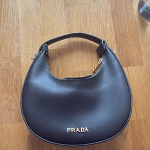 Svart handväska från Prada i skinn - Snygg svart handväska från Prada i rundad form med guldiga detaljer och dragkedja. Väskan har både ett kort handtag och en avtagbar axelrem, vilket gör den flexibel att bära. Tillverkad i slätt skinn och pryds av Pradas logga i guld framtill.