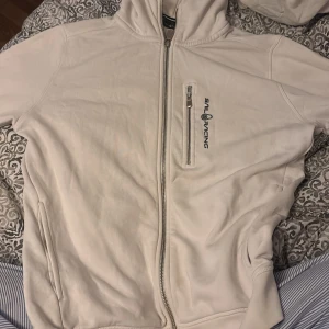 Vit hoodie från Sail Racing med dragkedja - Vit hoodie från Sail Racing med hel dragkedja framtill och en extra bröstficka med dragkedja. Tröjan har huva och långa ärmar, samt diskret logga på bröstet. Dock är det små fläckar vid ärmarna som jag inte har lyckats få bort. Och sliten i ärmarna ner till. Vilket man kan testa fixa osv. Priset kan även diskuteras pågrund av slitenhet.