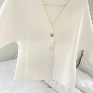 Vit v-ringad blus med knappar - Säljer en stilren vit blus med v-ringning och två dekorativa silvriga knappar framtill. Blusen har en rak, croppad passform och är tillverkad i ett mjukt, lätt ribbat material. Perfekt för dig som gillar minimalistisk och clean stil.