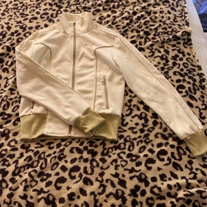 Vit ziptröja från Puma med ribbade muddar - Säljer en snygg vit ziptröja från Puma med höga ribbade muddar i beige och dragkedja framtill. Tröjan har två fickor med dragkedja och diskret Puma-logga på bröstet. Perfekt för en sportig look eller chill vardagsstil.