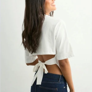 Vit croppad topp med knyt i ryggen - Säljer en vit croppad topp med korta ärmar och snygg öppen rygg med breda knytband. Toppen har en enkel och stilren design framtill och är tillverkad i mjuk bomull. Perfekt för dig som vill ha en trendig och luftig look.