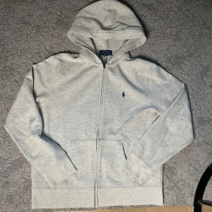 Polo Ralph Lauren zip up hoodie - Snygg ljusgrå hoodie från Polo Ralph Lauren med dragkedja och klassisk broderad logga på bröstet. Tröjan har huva, känguruficka och är tillverkad i mjuk bomullsmix. Perfekt för chill dagar eller när du vill ha en clean look. 14-16 yr 