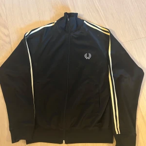 Svart Fred Perry track jacket S - Svart track jacket från Fred Perry med vita ränder längs ärmarna och klassisk broderad logga på bröstet. Jackan har hel dragkedja, ribbade muddar och hög krage. Perfekt för en sportig och stilren look.