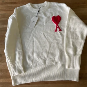 Vit stickad tröja från Ami - Vit stickad tröja från Ami med rund halsringning och ribbade muddar. På bröstet finns ett rött hjärta med bokstaven A. Perfekt för dig som gillar stilrena och snygga detaljer.