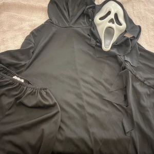 Halloween kostym  Scream - Svart kostym  Scream, perfekt för halloween eller temafest. Tröjan har långa ärmar och är tillverkad i ett mjukt syntetmaterial. Oversized passform för en dramatisk look. Har även byxor och tröja i samma serie och en dress med luva. Man får med allt på köpet även masken 