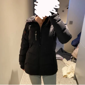 Svart pufferjacka från Sail Racing - Svart pufferjacka från Sail Racing med huva och dragkedja framtill. Jackan har en markerad logga på bröstet och en patch på ärmen. Flera fickor med dragkedja och en clean, sportig look. Perfekt för kalla dagar och riktigt snygg passform. Den ligger på värde runt 6000kr 