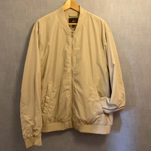 Beige bomberjacka från H&M XXL - Snygg beige bomberjacka från H&M i storlek XXL. Jackan har klassisk bomberkrage, dragkedja framtill och två snedställda fickor. Tillverkad i ett lätt syntetmaterial med ribbade muddar vid ärmslut och nederkant. Perfekt för dig som gillar stilren och enkel look.