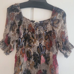 Blommig blus från Odd Molly - Supersöt blus från Odd Molly i luftig viskos med blommigt och prickigt mönster i beige, blått, rött, orange och rosa. Blusen har korta puffärmar med resår och smockdetaljer samt en lös passform. Perfekt för dig som gillar bohemisk och färgglad stil.