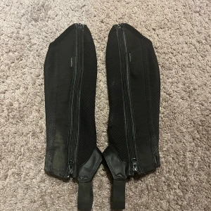 Svarta ridchaps från Fouganza - Säljer ett par svarta ridchaps från Fouganza. De är tillverkade i en kombination av skinn och syntetmaterial, med dragkedja längs sidan och elastiska band nedtill. Dock lite skavanker då de är mycket använda.