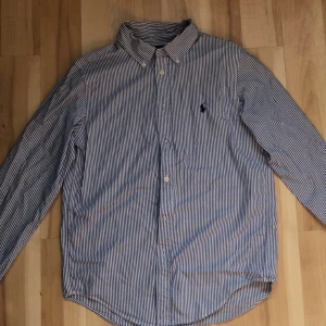 Blårandig skjorta från Ralph Lauren - Snygg blårandig skjorta från Ralph Lauren med klassisk krage och broderad logga på bröstet. Skjortan har lång ärm, knappar framtill och är gjord i mjuk bomull. Passformen är custom fit vilket ger en schysst siluett. Perfekt för dig som gillar stilrena och tidlösa plagg. Storlek s