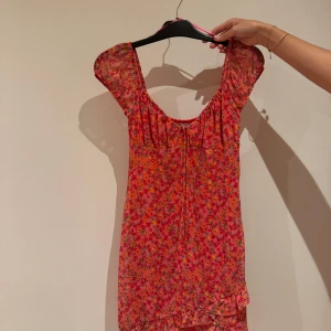 Blommig kortklänning från Bershka - Söt kortklänning från Bershka i rosa och rött med blommigt mönster. Klänningen har korta puffärmar, rundad halsringning och volangdetalj längst ner. Perfekt för sommarens alla tillfällen, lätt och luftig i materialet.