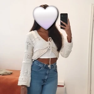 Vit croppad stickad kofta med snörning - Superfin vit croppad stickad kofta med snörning framtill och v-ringad hals. Koftan har långa ärmar och ett snyggt hålmönster som ger en boho-vibe. Perfekt att slänga över ett linne eller topp för en chill look.