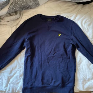 Mörkblå sweatshirt från Lyle & Scott - Mörkblå sweatshirt från Lyle & Scott med klassisk rund hals och broderad gul logga på bröstet. Tröjan har långa ärmar och ribbade muddar vid ärmslut och nederkant. Perfekt för en stilren och avslappnad look.