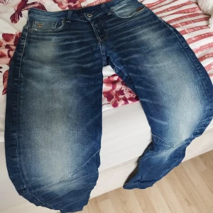 Blå G-Star ARC jeans med tvättad look - Säljer ett par blå G-Star ARC jeans med snyggt tvättad effekt och markerade slitningar. Jeansen har klassisk femficksdesign, silverfärgade detaljer och logga på fickan. Modellen har en avslappnad passform och är tillverkad i robust denim.