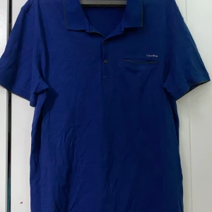 Blå Calvin Klein t-shirt slim fit - Snygg marinblå t-shirt från Calvin Klein i slim fit-modell. T-shirten har korta ärmar, krage med knappslå och en liten bröstficka med diskret Calvin Klein-logga. Perfekt för dig som gillar stilrena och klassiska plagg.