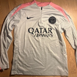 PSG långärmad träningströja Nike - Snygg långärmad fotbollströja från Paris Saint-Germain med halv dragkedja. Tröjan är ljusgrå med rosa axelpartier, svart Nike-logga och PSG-märke på bröstet. Qatar Airways som sponsortryck framtill. Perfekt för träning eller match.