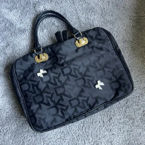 Svart datorväska från DKNY - Väldigt snygg svart datorväska från DKNY med tydligt logomönster över hela väskan. Väskan har två handtag i skinn, guldfärgade metalldetaljer. (Rosetterna tillkommer inte) Ja, väskan får plats med dator såklart.
