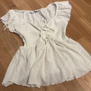 topp - Supersöt vit blus med knyt framtill och volangiga korta ärmar. Tillverkad i ett lätt, krinklat bomullstyg som ger en luftig känsla. Perfekt för varma dagar och ger en romantisk vibe till din outfit.